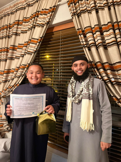 A Madrassa An-Nur wal-Huda student receiving a certificate of attendance and participation from his teacher